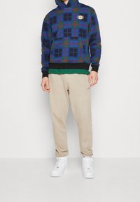 La sudadera con capucha presenta un diseño de cuadros en azul y verde con acentos en negro; se combina con pantalones de chándal beige y zapatillas altas blancas.