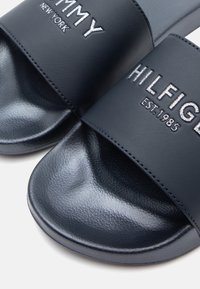 Navy blue slide sandals made of smooth synthetic material, featuring embroidered logos, rounded edges, and a textured footbed for grip.