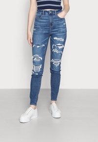 Högmidjade blå denimjeans med slitna hål och fransar längs låren, i kombination med vita sneakers. Mjuk textur, åtsittande design.
