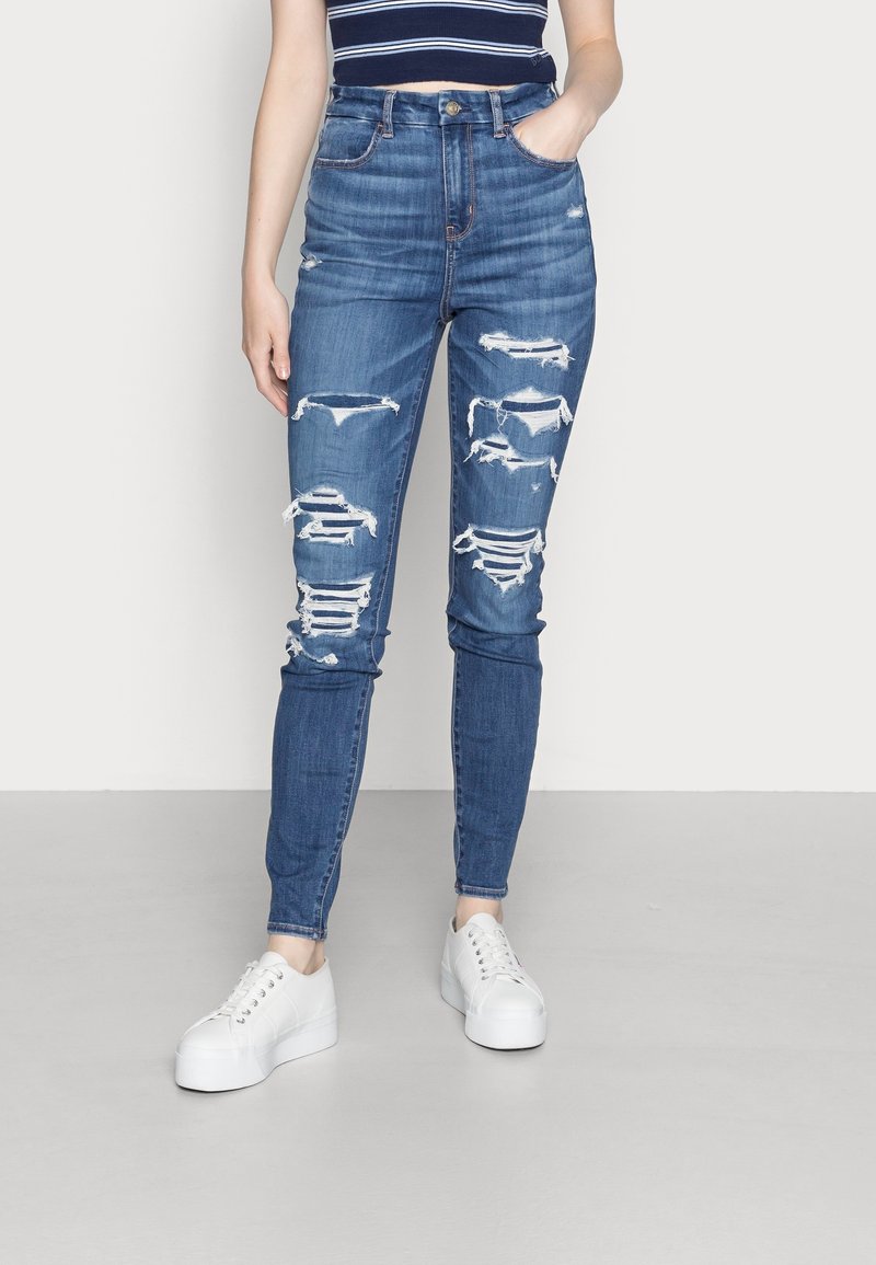 Högmidjade blå denimjeans med slitna hål och fransar längs låren, i kombination med vita sneakers. Mjuk textur, åtsittande design.
