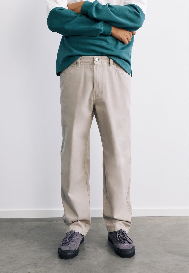 Personne debout, bras croisés, portant un pantalon beige, un sweat-shirt bleu sarcelle et des baskets grises devant un mur blanc et un sol gris.