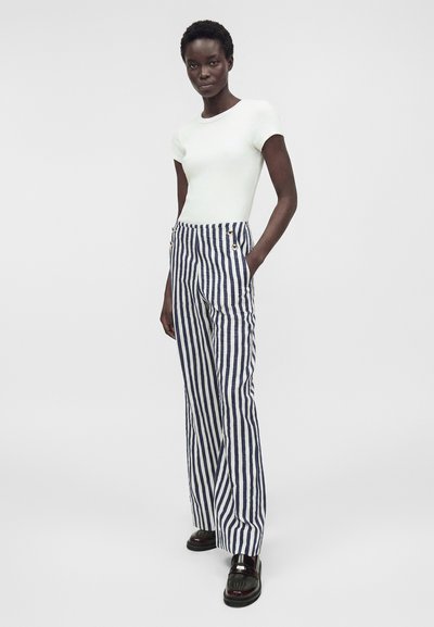 Γυναίκα που φοράει λευκή εφαρμοστή μπλούζα με κοντά μανίκια, παντελόνι σε navy και λευκές κάθετες ρίγες με κουμπιά στη μέση και μαύρα loafers, στέκεται με το χέρι στην τσέπη.