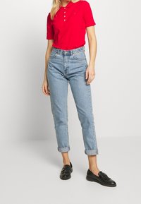 Chemise polo rouge à manches courtes et trois boutons, associée à un jean taille haute bleu clair retroussé aux poignets et des mocassins noirs.