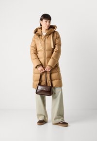 Brauner Puffer-Mantel mit Kapuze, Reißverschluss und auffälligen gestickten Details; kombiniert mit hellblauen, weit geschnittenen Hosen und einer braunen Handtasche.