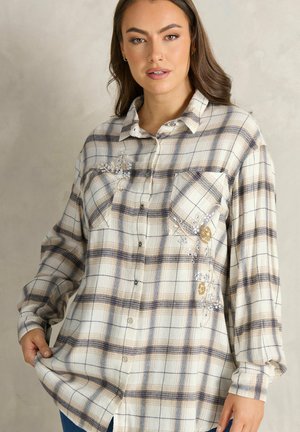 Kvinde iført en beige og blå ternet knap-op-skjorte med blomsterbroderi på brystlommerne, holder skjortens kant og står foran en ensfarvet baggrund.