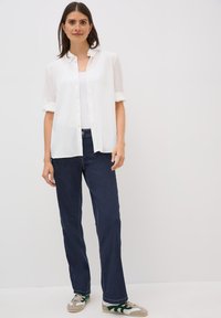Chemise blanche à boutons, texturée, avec des manches retroussées sur un débardeur blanc, associée à un jean droit bleu foncé et des baskets grises.