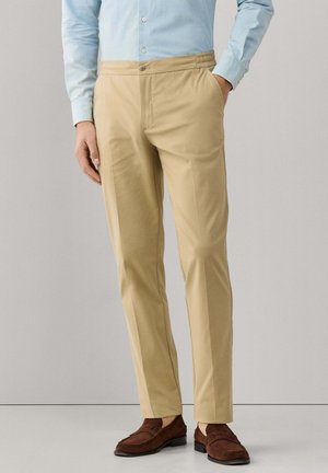 Man draagt beige op maat gemaakte broek, lichtblauw overhemd en bruine suède loafers, staand met één hand in de zak tegen een effen achtergrond.