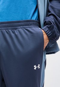 Άτομο που φορά μπλε αθλητικό σορτς με λευκό λογότυπο Under Armour, μπλε μπλουζάκι και μπουφάν, με το χέρι στην τσέπη του σορτς.