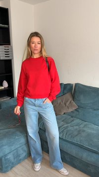 Rød bomullsgenser, lyseblå denimjeans, står på en blågrønn kordfløyelssofa. Holder røde solbriller, med langt hår og minimale tilbehør.