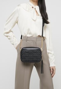 Musta nahkainen olkalaukku, jossa tikattu kuvio, kultainen ketjuhihna ja hapsukoriste, jota kantaa henkilö luonnonväreissä pukeutuneena.