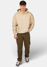 Felpa oversized beige con logo bianco, tasca frontale e cordoncino, abbinata a pantaloni in corduroy marroni e sneakers bianco e nere.