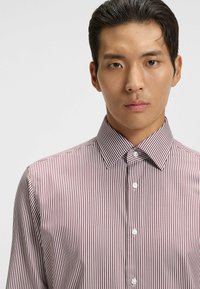 Jeune homme aux cheveux courts et foncés portant une chemise blanche avec de fines rayures verticales bordeaux et un col boutonné.