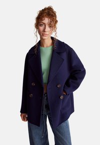 Cappotto peacoat in lana navy con chiusura a doppio petto, ampi revers e tasche laterali. Indossato sopra un top verde corto e jeans a vita alta.