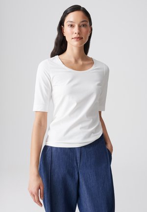 Vrouw met lang donker haar, gekleed in een wit topje met korte mouwen en blauwe wijde broek, staand tegen een eenvoudige lichte achtergrond.