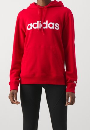 Osoba v červené mikině Adidas s bílým logem a černých legínách stojí před jednobarevným světlým pozadím.