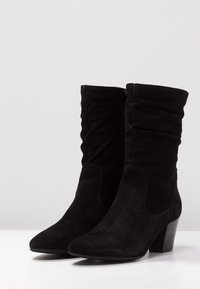 Une paire de bottes noires en daim mi-mollet avec des orteils pointus, des tiges froncées, des fermetures éclair sur le côté et des talons carrés sur une surface blanche.