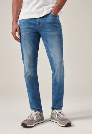 Man som bär ljusblå slim-fit jeans, vit t-shirt och beige sneakers, stående med ena handen i fickan mot en ljus bakgrund.