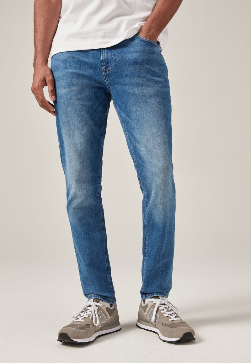 Mies, joka käyttää vaaleansinisiä slim fit -farkkuja, valkoista t-paitaa ja beigejä lenkkareita, seisoo yhdellä kädellä taskussa vaalealla taustalla.