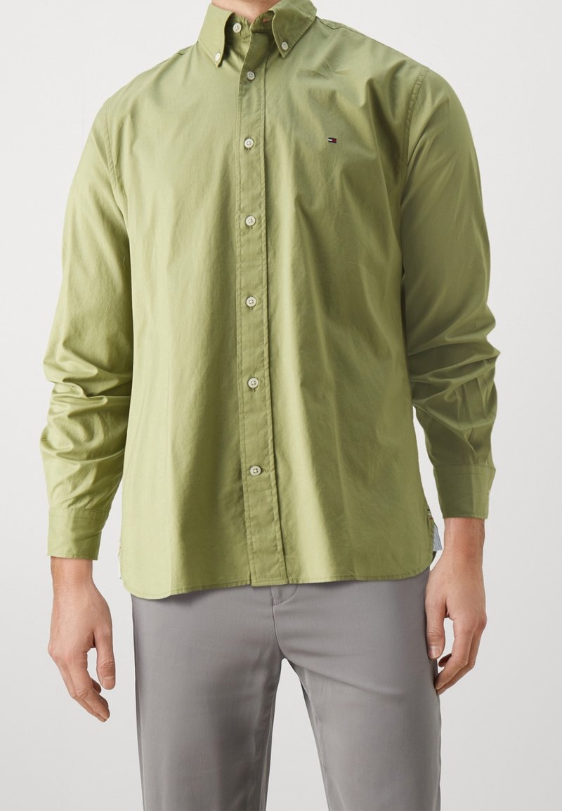 Homme portant une chemise à manches longues olive boutonnée et un pantalon gris, debout les bras détendus le long du corps sur fond blanc.