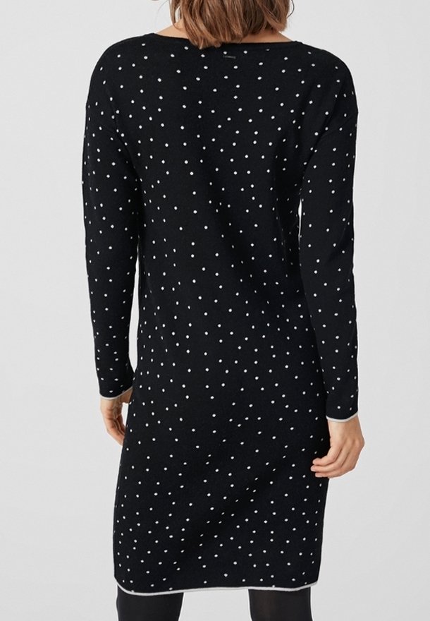 Femme portant une robe noire longue aux manches longues, longueur genou, à pois blancs, vue de dos sur un fond clair uni.