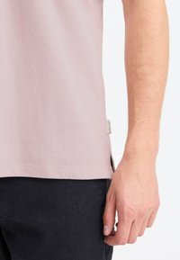 Camisa de manga corta de color rosa claro con tejido texturizado y aberturas laterales. Presenta una etiqueta en el dobladillo inferior. Combinada con pantalones de color oscuro.