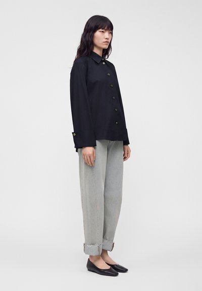 Femme debout portant une chemise noire boutonnée, un jean gris clair retroussé et des chaussures plates noires sur un fond uni gris clair.