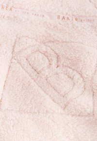 Miękki różowy koc wykonany z teksturowanej tkaniny, z wypukłym logo 'b' na środku i subtelną etykietą marki wzdłuż krawędzi.