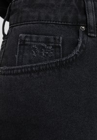 Gros plan d’un jean en denim noir montrant la poche avant, la petite poche avec une usure subtile, les coutures et un détail de rivet.