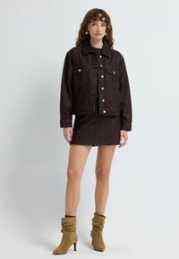 Vrouw in een donkerbruine denim jack en bijpassende mini-jurk met tan suède enkellaarsjes, staand tegen een effen lichte achtergrond.