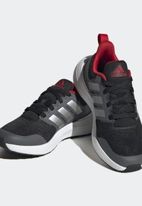 Zapatillas deportivas en negro y gris con detalles en rojo, parte superior de malla, tres franjas plateadas, cuello acolchado y suela de goma blanca.
