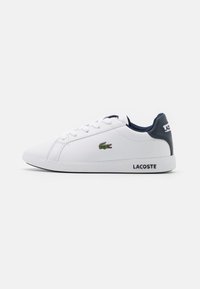 White synthetic sneakers with navy accents, lace-up design, and embroidered crocodile logo. Flat sole with "LACOSTE" branding on the side.