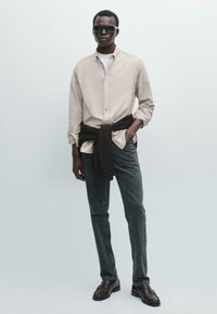 Camicia beige con maniche arrotolate, jeans scuri e mocassini marroni. Un maglione nero è annodato in vita. Gli occhiali da sole completano il look.