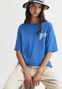 T-shirt en coton bleu à manches courtes et col rond, avec un petit graphique de motifs de fruits sur la poitrine. Pantalon beige clair.