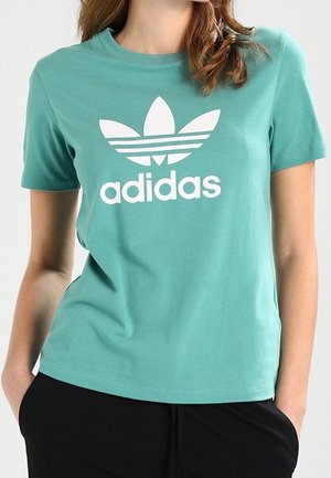Femme portant un t-shirt Adidas turquoise avec un logo trèfle blanc et du texte, associé à un pantalon noir, mains dans les poches.