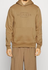 Brown hoodie with a front pocket, drawstring hood, and "HILFIGER" logo. Soft texture, ribbed cuffs, and hem, made of sweatshirt fabric.