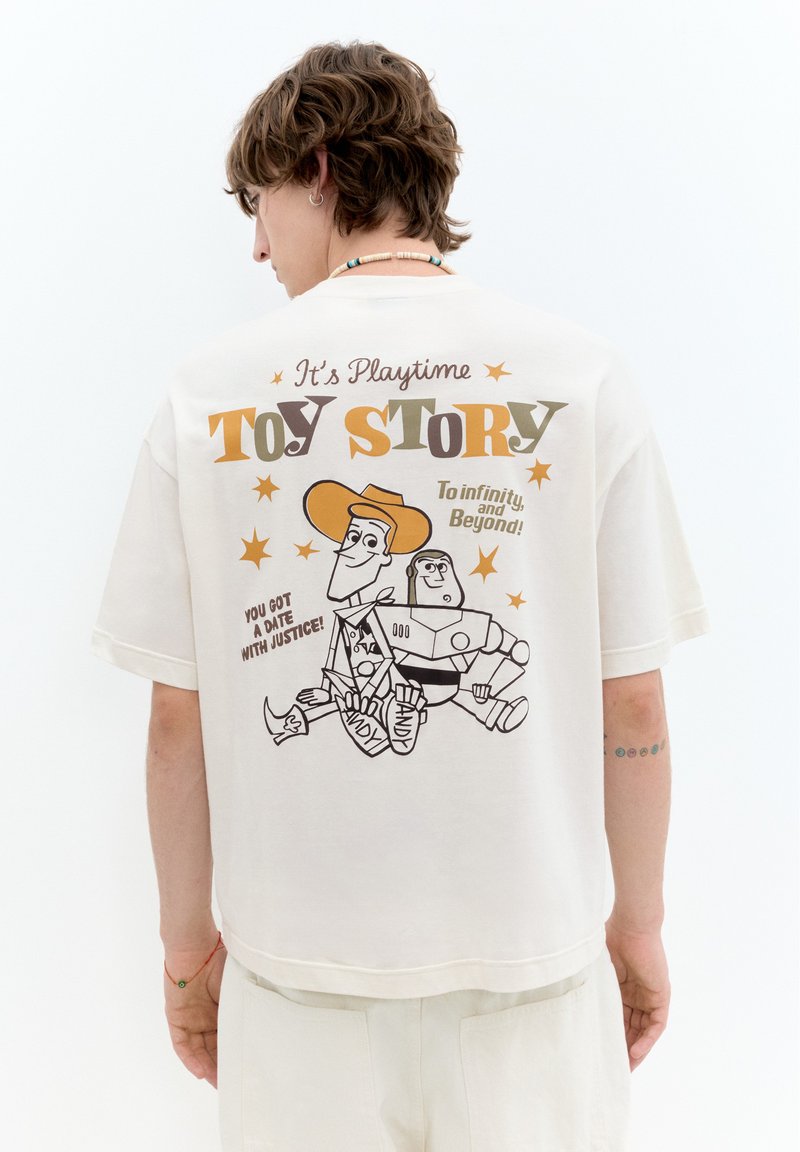 Personne portant un t-shirt blanc Toy Story représentant les personnages de dessin animé Woody et Buzz Lightyear avec les phrases "It's playtime" et "To infinity and beyond."
