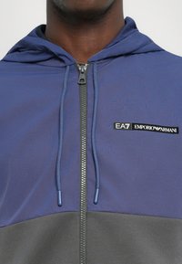 Plava i siva zip-up dukserica s kapuljačom, koja ima teksturiranu tkaninu, podesive vezice i EA7 Emporio Armani logo na prsima.
