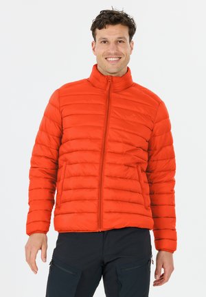 Man glimlacht, staat, draagt een feloranje donsjas met ritssluiting aan de voorkant en een zwarte broek, tegen een lichte effen achtergrond.