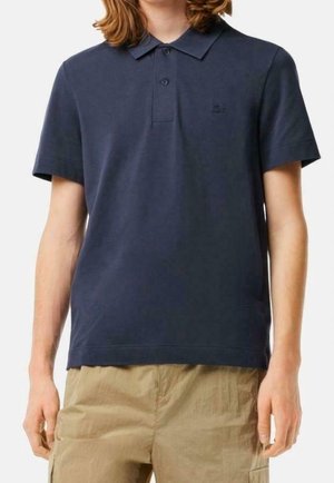 Person trägt ein dunkelblaues Poloshirt mit kurzen Ärmeln und hellbraune Cargohosen, steht mit entspannten Armen an den Seiten.