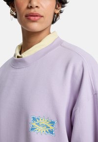 Μωβ φούτερ με ριμπ λαιμόκοψη, φορεμένο πάνω από ένα ανοιχτό κίτρινο πουκάμισο, με πολύχρωμο γραφικό λογότυπο στο στήθος.