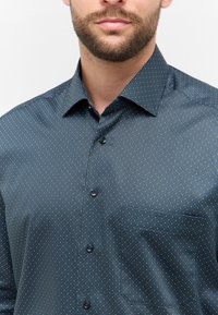 Camicia blu navy con un piccolo motivo geometrico, realizzata in un tessuto morbido. Presenta un colletto classico e una tasca sul petto.