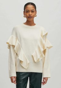 Crème trui met asymmetrische ruffle-accenten, zachte textuur en geribbelde manchetten, gecombineerd met donker groene leren broek.