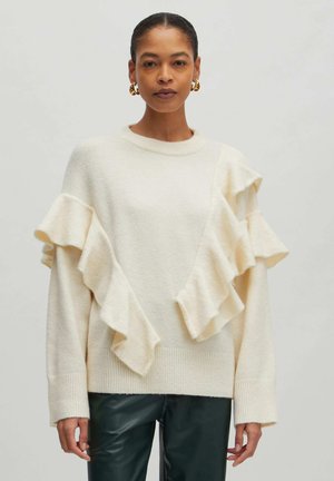 Crème trui met asymmetrische ruffle-accenten, zachte textuur en geribbelde manchetten, gecombineerd met donker groene leren broek.