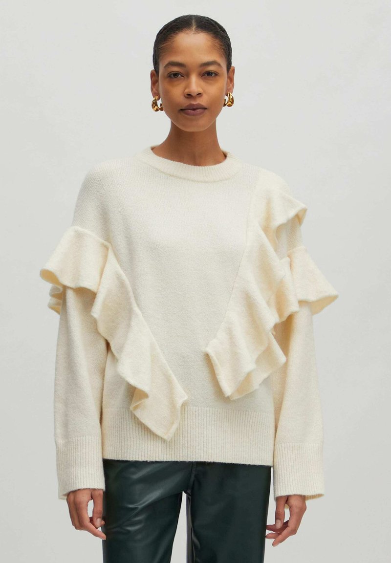 Crème trui met asymmetrische ruffle-accenten, zachte textuur en geribbelde manchetten, gecombineerd met donker groene leren broek.