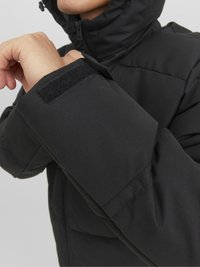 Giacca nera imbottita con colletto alto, chiusura a zip e tessuto testurizzato, dotata di dettaglio con strap in Velcro sul polsino.