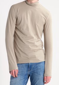Beige Langarmrollkragenoberteil aus glattem Stoff, mit einer taillierten Passform und verlängerten Bündchen, kombiniert mit blauen Jeans.