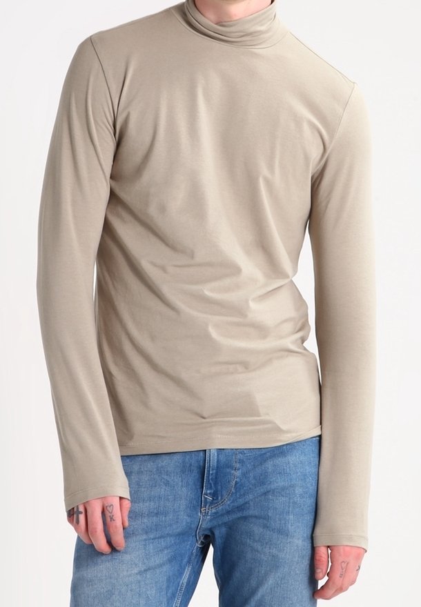 Beige Langarmrollkragenoberteil aus glattem Stoff, mit einer taillierten Passform und verlängerten Bündchen, kombiniert mit blauen Jeans.