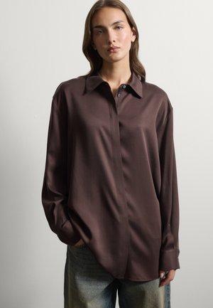 Jonge vrouw met steil haar, die een losse donkerbruine blouse met knopen en lichtblauwe spijkerbroek draagt, staand tegen een eenvoudige lichte achtergrond.
