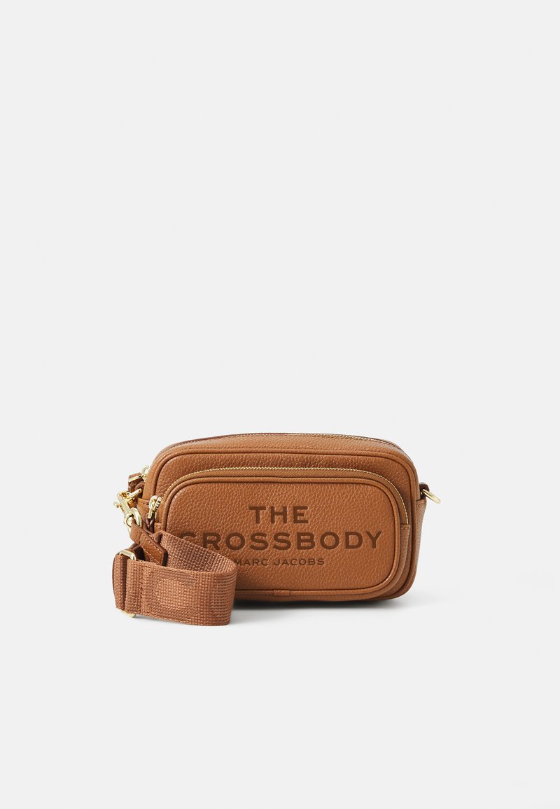 Ruskea nahkainen olkalaukku, jossa on kultaiset yksityiskohdat, kohokuvioitu teksti "THE CROSSBODY", suorakulmainen muoto ja säädettävä kankainen olkahihna.