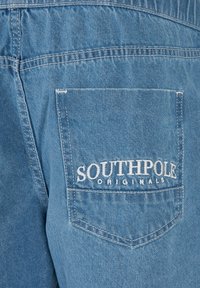 Poche arrière d'un jean denim bleu avec un texte brodé blanc "SOUTHPOLE ORIGINALS" sur la poche.